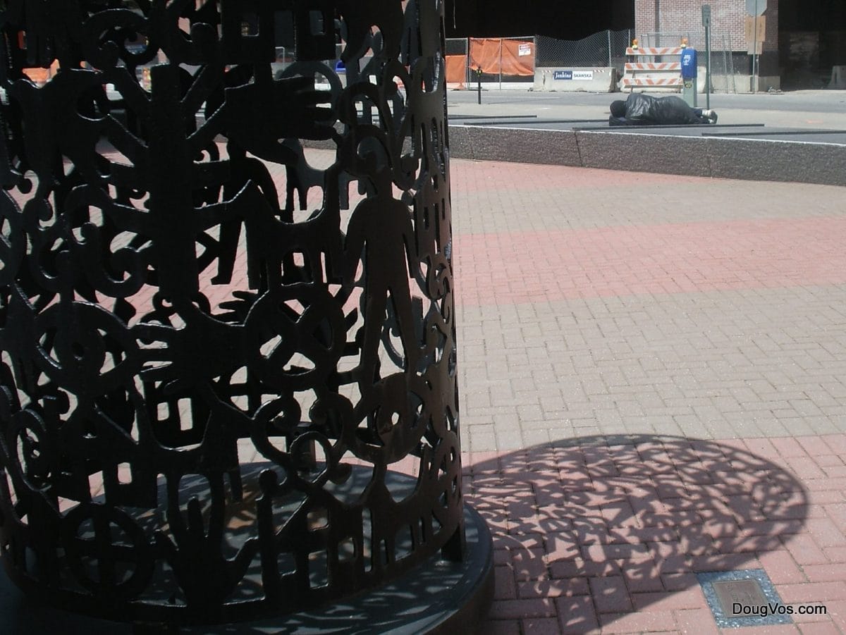 Foreground: Iron hands sculpture in downtown Detroit. Background: Street person is sleeping on some iron grates.