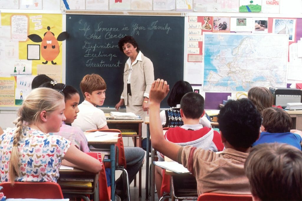 Lessons On Happiness From The School of Life. Teacher in a classroom writes something on the blackboard - If someone in your family has cancer - how does that make you feel?