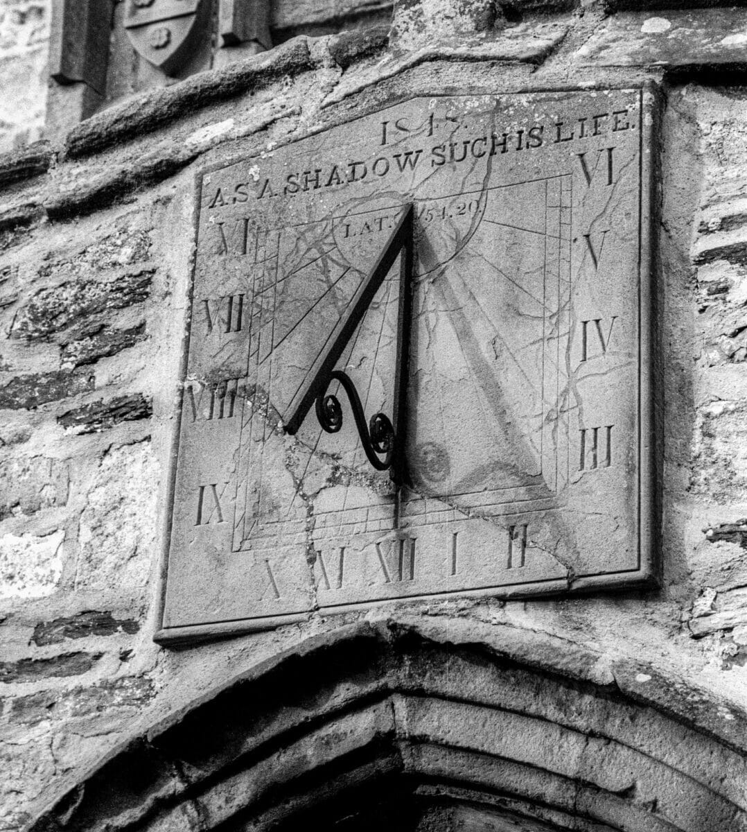 Sundial shadows are revealed on an old sundial placed above an archway, dated 1848. Roman numerals are inscribed to indicate the hours of the day. The words inscribed at the top say: As a shadow, such is life.