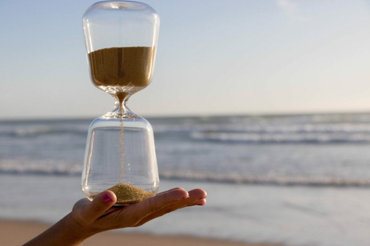 A hand holding an hourglass, with ocean and beach in the background. Sands of time photography.