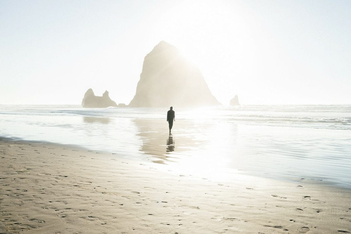 A silhouette of a man walking through the shallow water on a sandy beach.